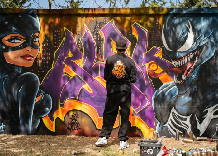 A street artist paints a colorful superhero-themed graffiti on an outdoor wall, featuring vivid characters.